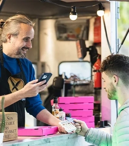 Ein Mitarbeiter eines Imbisswagens in einer Schürze hält ein Smartphone und interagiert mit einem Kunden, der ein Zahlungsterminal hält, umgeben von rosa Kartons und Marktartikeln.