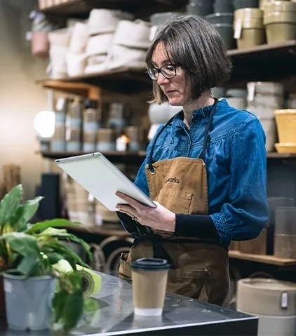 Eine Person mit Brille, blauem Hemd und Schürze hält ein Tablet in einem Geschäft oder Werkstatt mit einer Topfpflanze und einem Kaffeebecher auf dem Tisch und Regalen mit Behältern im Hintergrund.